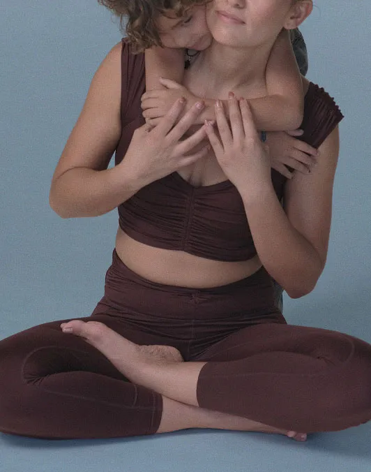 Woman in burgundy activewear sitting cross-legged while a child hugs her from behind.